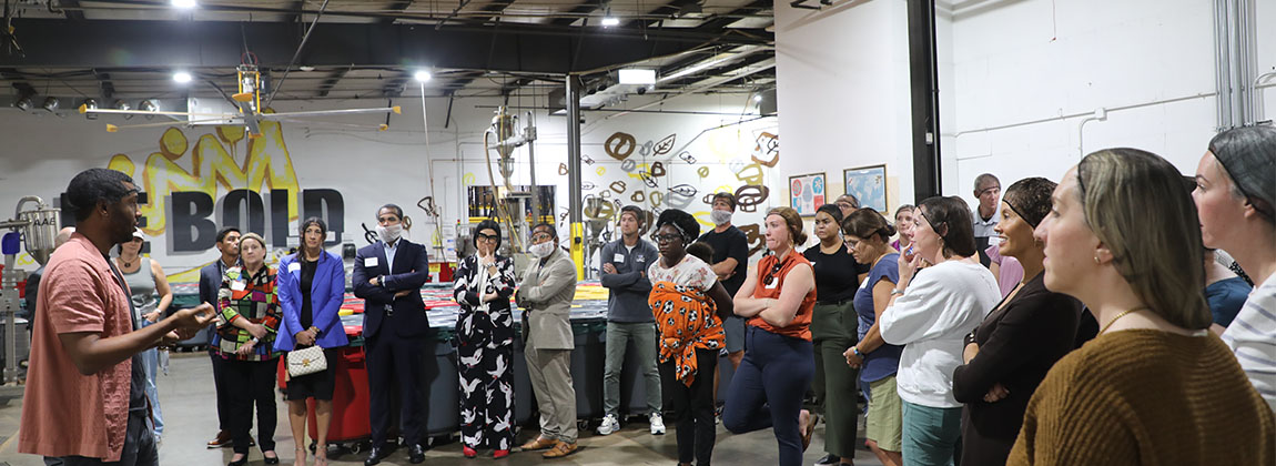 A group of people standing in a factory listening to a man speak.